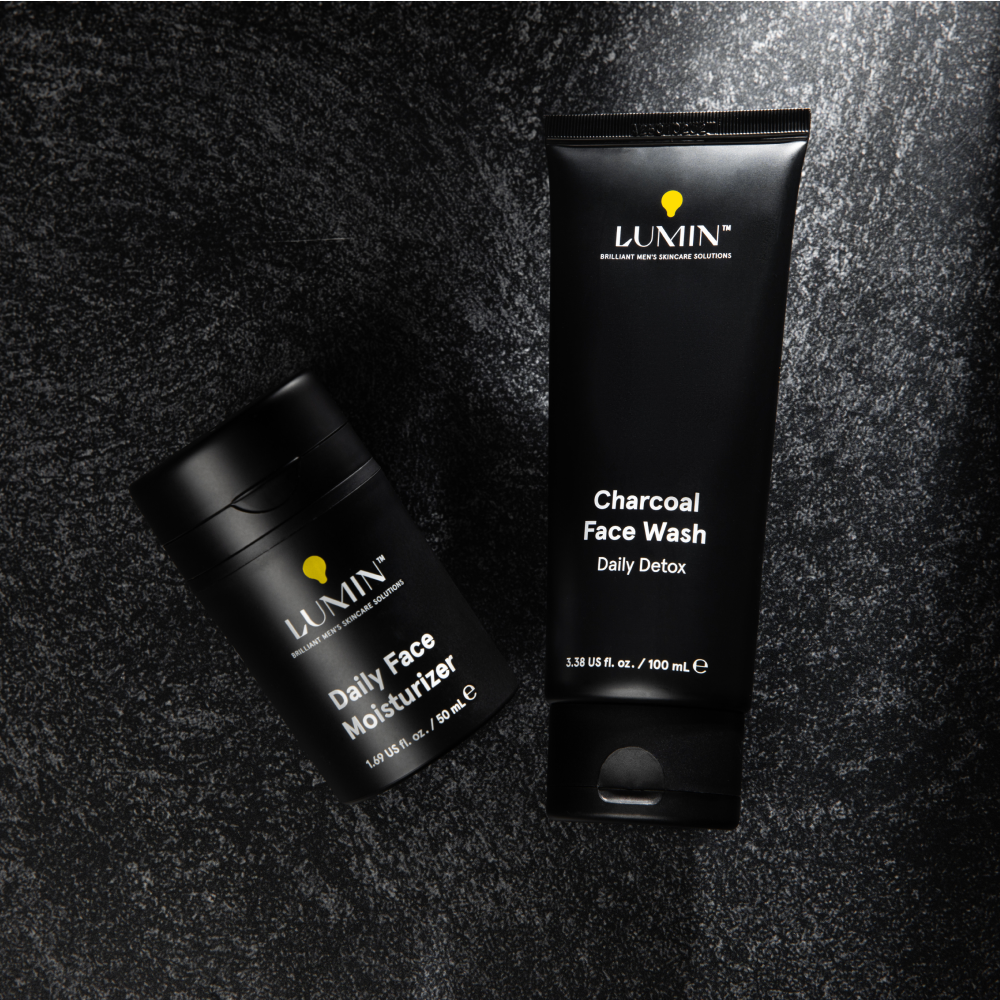 Lumin men's skincare charcoal face wash and daily face moisturizer on textured black surface for daily detox and hydration.