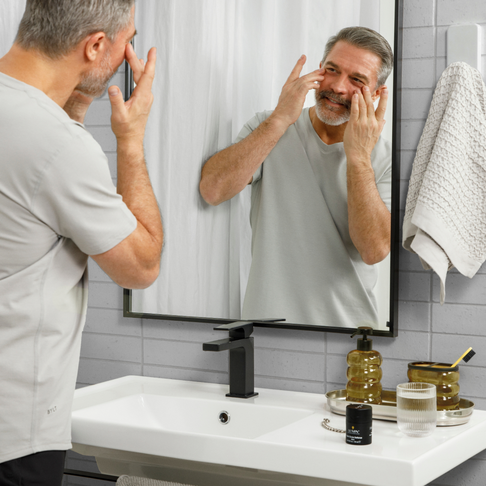 Middle-aged man applying facial skincare cream in modern bathroom with mirror, black faucet, and toiletry set on sink.