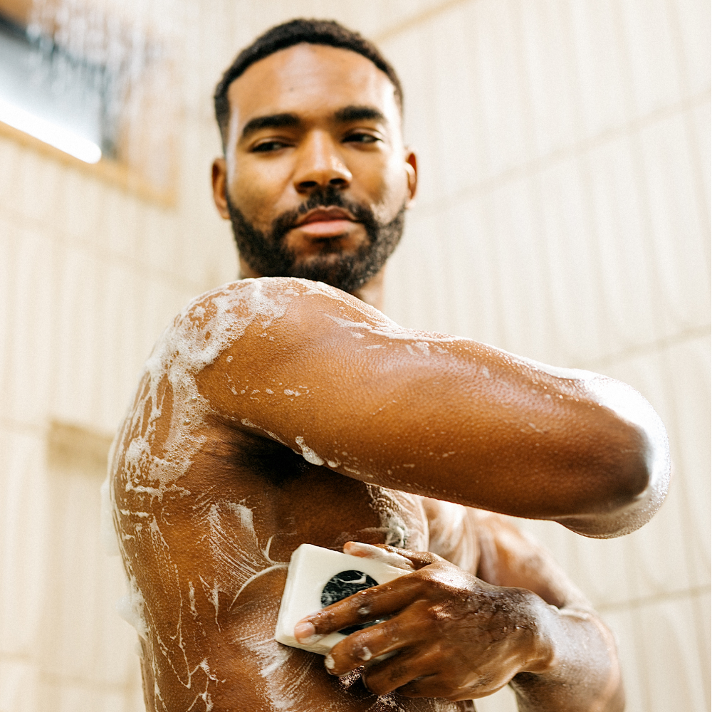 Bearded man washing his upper body with white soap bar, covered in lather, in a tiled shower setting.