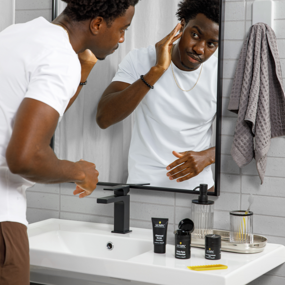 Man in white t-shirt grooming skin in bathroom mirror with skincare products, towel, and comb on sink counter