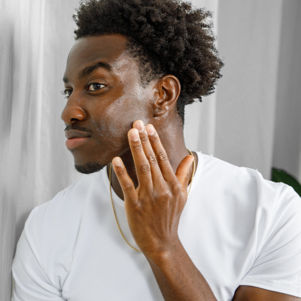 Young Black man applying moisturizing cream to face with gold chain, white shirt, curly hair, skincare routine for dry skin care