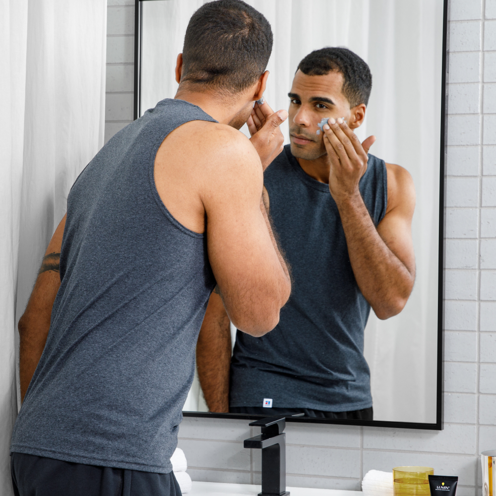 Man in sleeveless gray shirt applying blue face cream in bathroom mirror, modern white tiled wall, skincare routine essentials visible