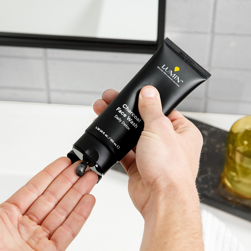 Lumin Charcoal Face Wash tube dispensing black daily detox cleanser onto hand over bathroom sink and mirror.
