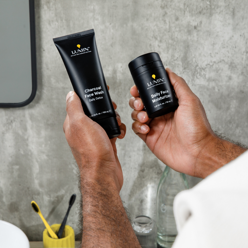 Lumin charcoal face wash and daily face moisturizer held by man against concrete bathroom wall with toothbrushes.