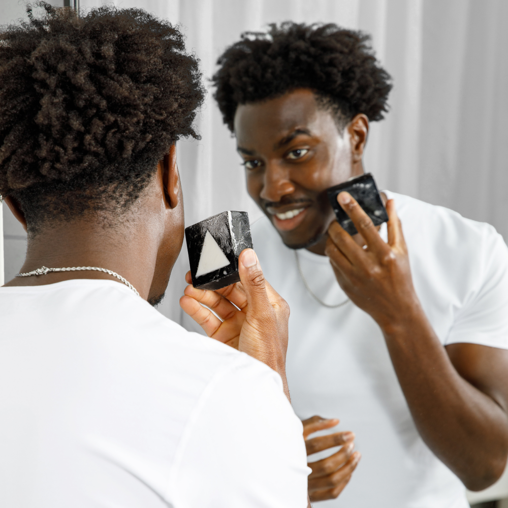 Young man with curly hair applying black and white facial soap bar, skincare routine in front of bathroom mirror.