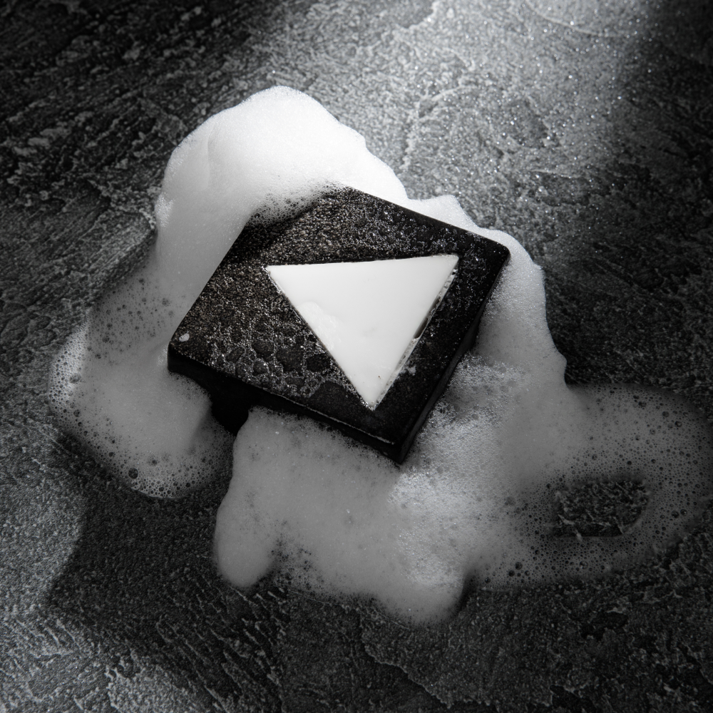 Black triangular bar soap with white triangle detail surrounded by white foam bubbles on textured dark surface