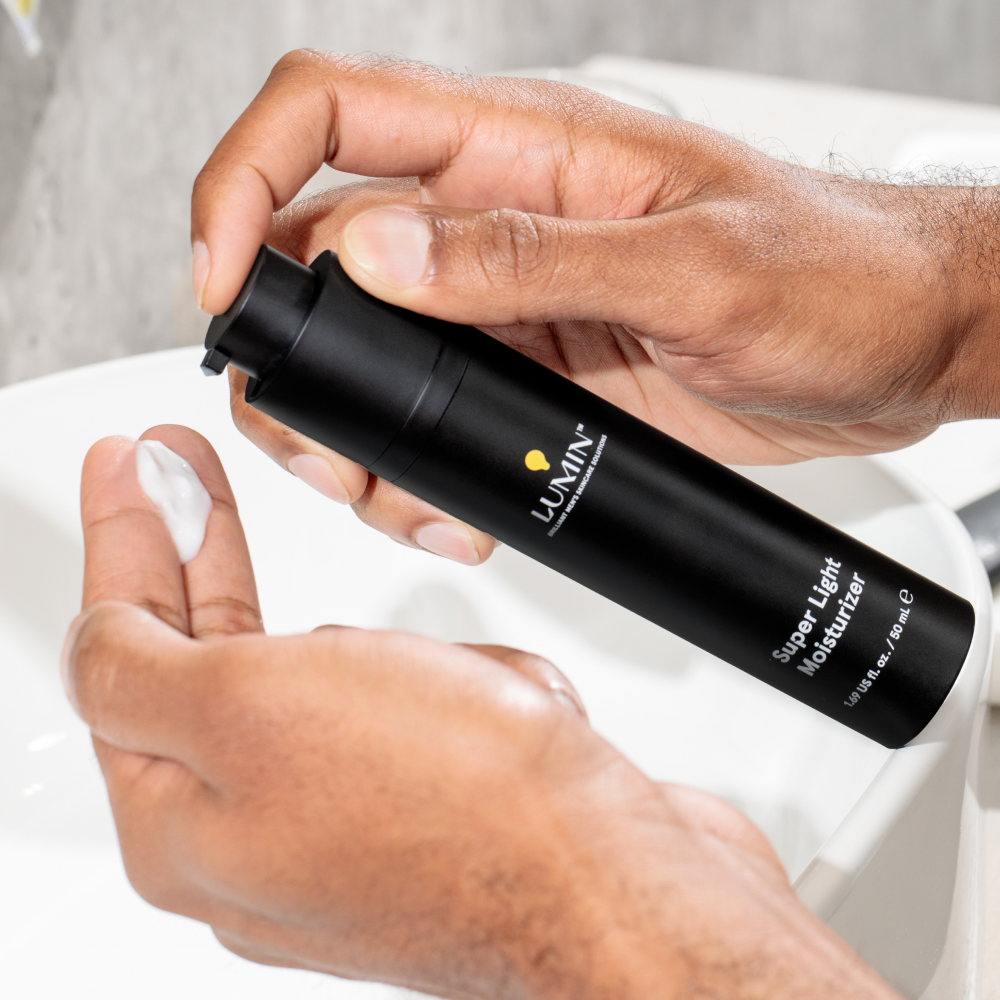 Hands dispensing Lumini Super Light Moisturizer cream from black pump bottle over white bathroom sink for skincare.