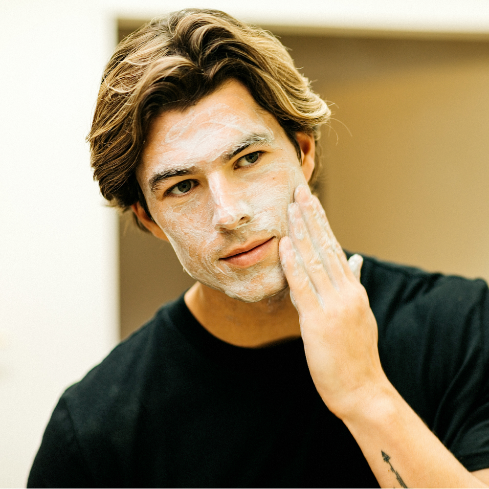 Young man with light brown hair applying white facial cleanser cream, skincare routine, grooming and self-care concept