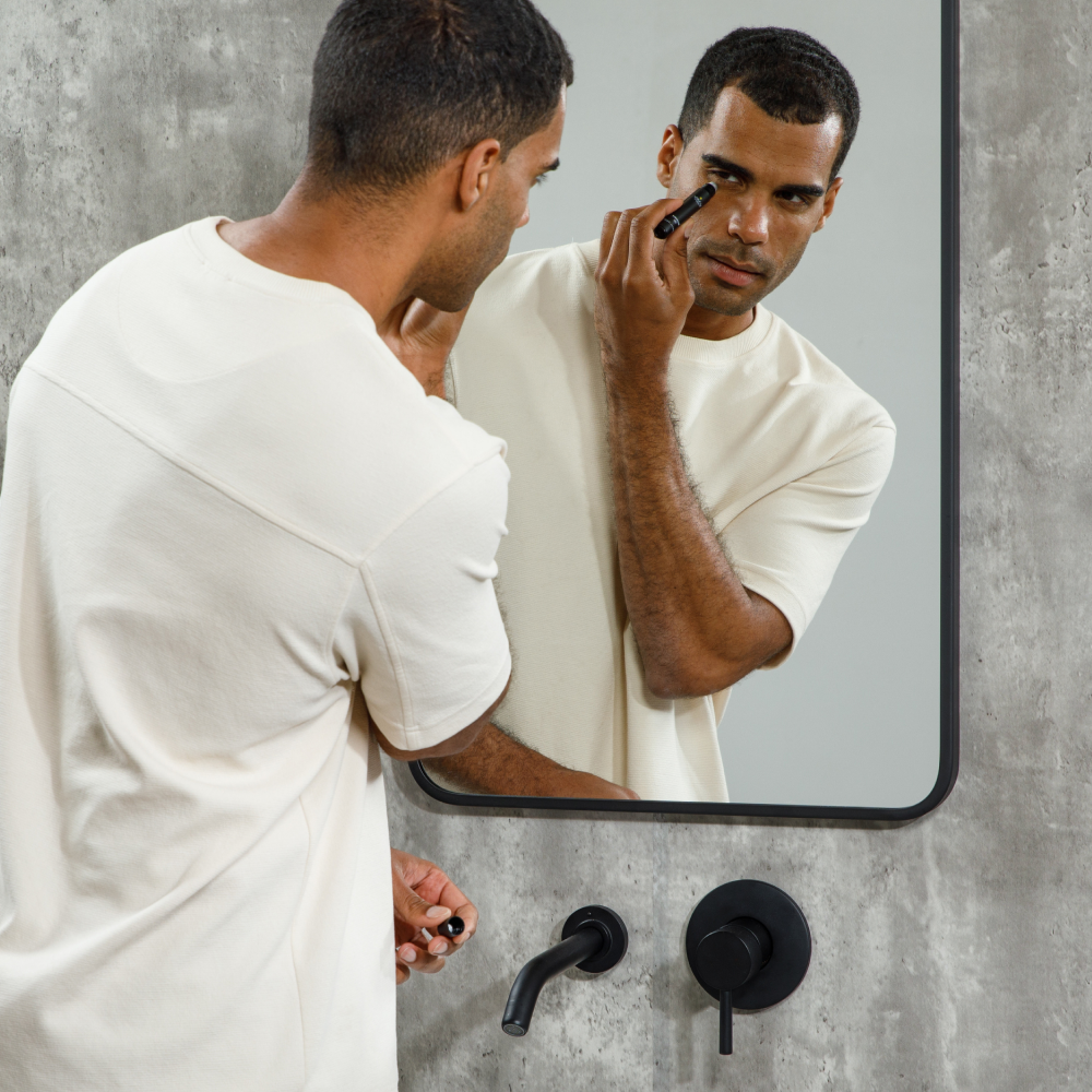 Man applying under-eye concealer with makeup brush in modern bathroom with concrete wall and black faucet.