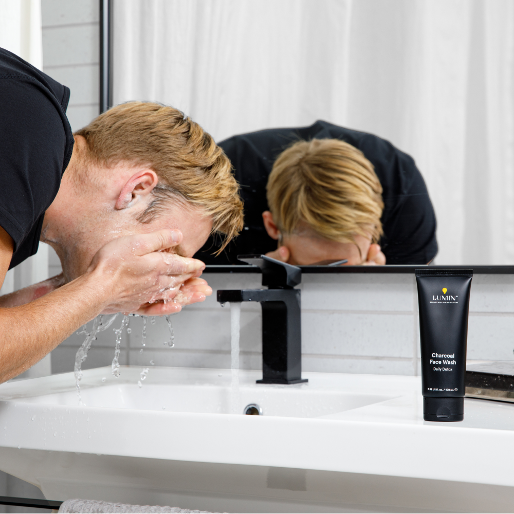 Man washing face with water at bathroom sink next to Lumin charcoal face wash tube and black faucet