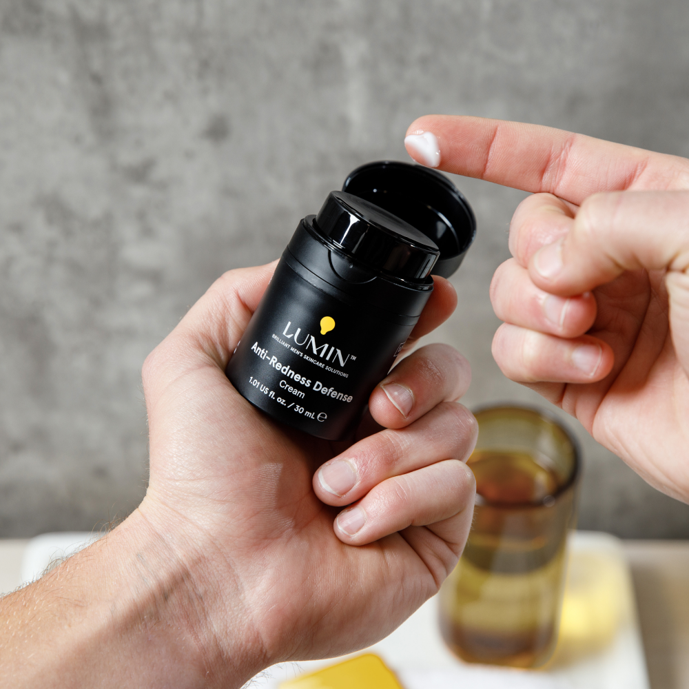 Black Lumini Anti-Redness Defense Cream container held with a dab of white cream on a finger, men's skincare product.