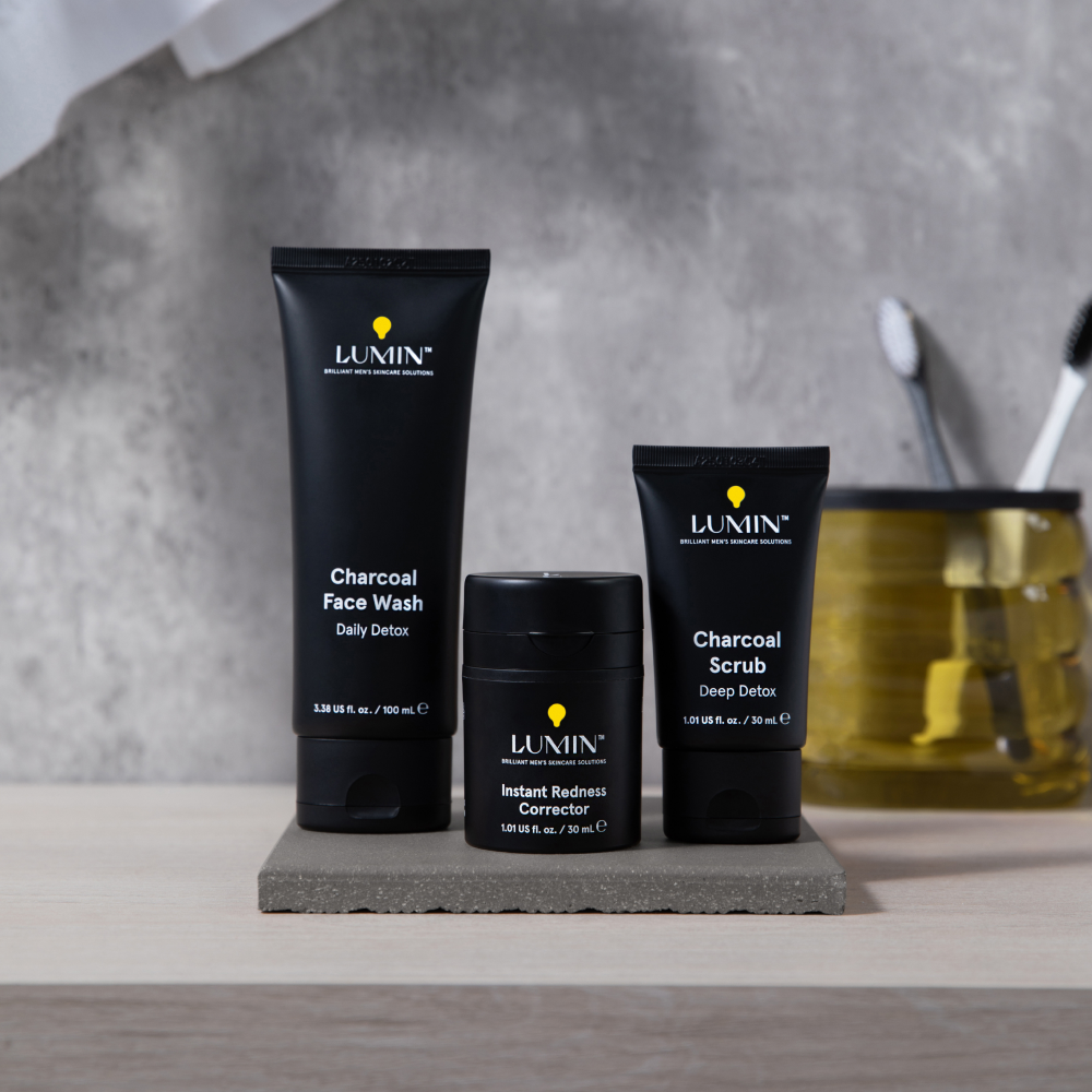 Lumin men's charcoal face wash, charcoal scrub, and instant redness corrector skincare products on gray stone tray with toothbrushes.