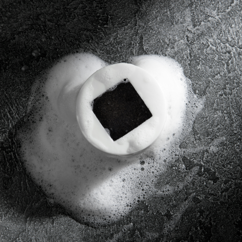 Black charcoal soap bar surrounded by white creamy foam on textured dark gray surface, skincare cleansing product.
