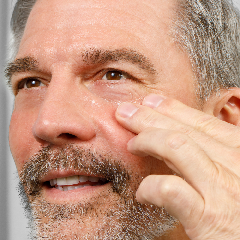 Middle-aged man applying eye cream under brown eyes, showcasing skincare for mature skin and anti-aging routine.