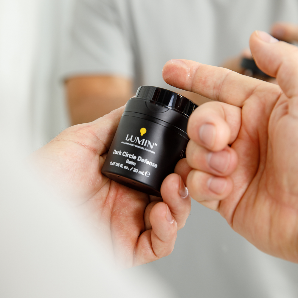Lumin Dark Circle Defense Balm container held in hand with cream on fingertip, skincare for men, anti-dark circles.
