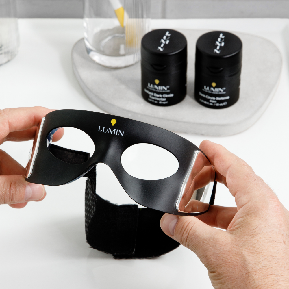 Lumin LED light therapy eye mask held by hands with two Lumin skincare jars on marble tray in background.